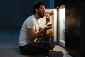 a young man in need of binge eating disorder treatment sits on the floor in front of a refrigerator eating